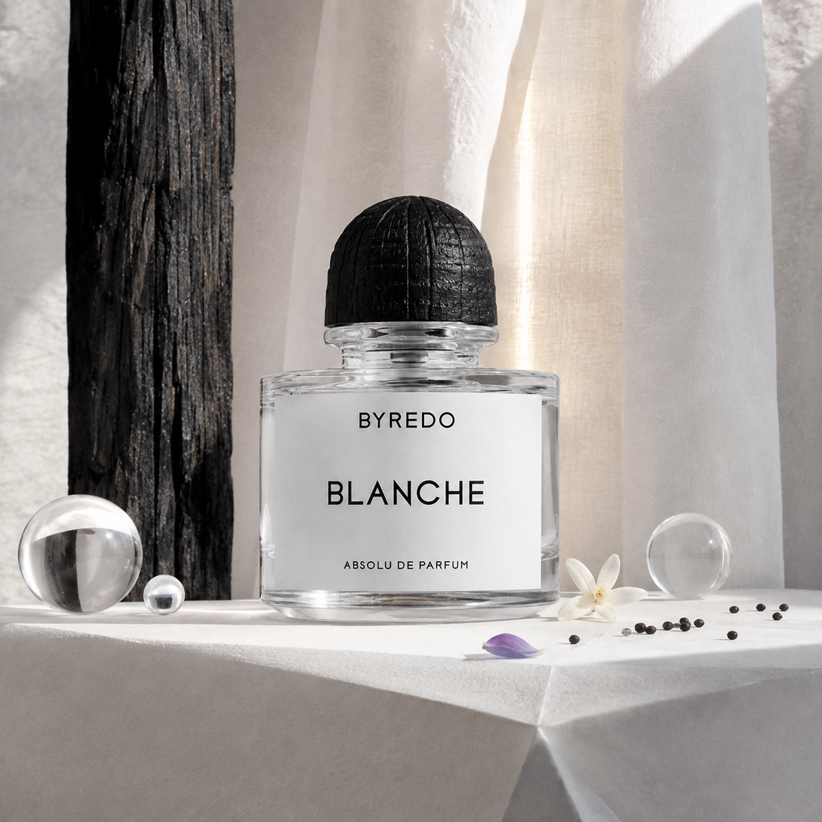 Byredo Blanche Absolu na bílém stole u závěsu se skleněnými koulemi a bílým květem – MrRizz